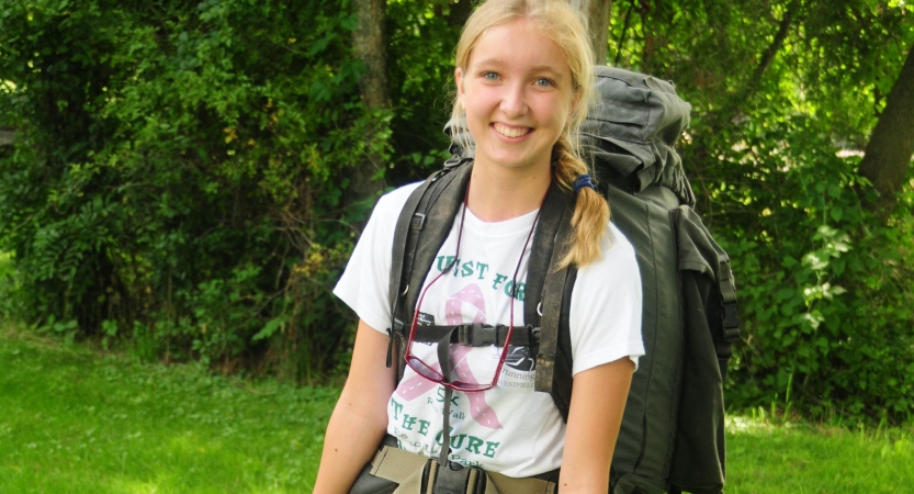 A person wearing a backpack smiles for the photo in a green wooded area. 